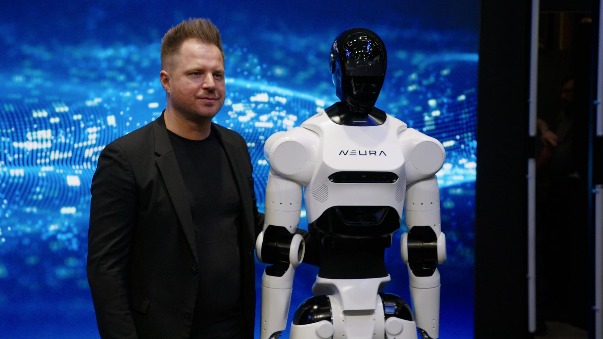 "WISO-Dokumentation: Junge Startups - Deutsche Hightech für die Weltspitze": David Reger steht im schwarzen Anzug vor einer blauen Animation auf einer Videowall. Neben ihm steht ein schwarz-weißer, lebensgroßer, humanoider Roboter.