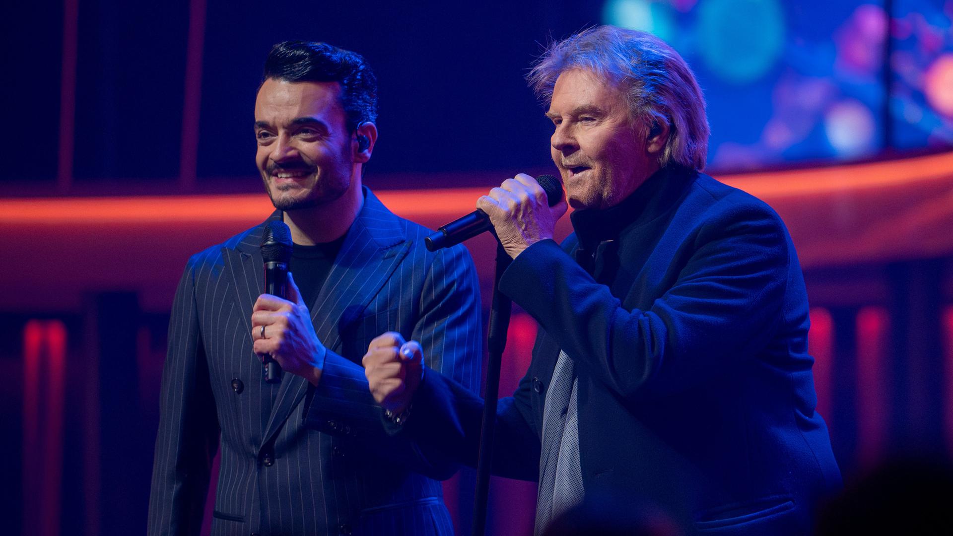 "Die Giovanni Zarrella Show - Eine Samstagnacht mit Howard Carpendale": Giovanni Zarrella und Howard Carpendale stehen nebeneinander auf der Bühne, halten Mikrofone in der Hand und lächeln. Beide tragen dunkle Anzüge. Der Hintergrund ist bunt beleuchtet und mit dekorativen Elementen versehen.
