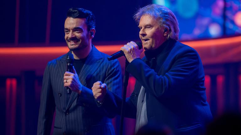 "Die Giovanni Zarrella Show - Eine Samstagnacht mit Howard Carpendale": Giovanni Zarrella und Howard Carpendale stehen nebeneinander auf der Bühne, halten Mikrofone in der Hand und lächeln. Beide tragen dunkle Anzüge. Der Hintergrund ist bunt beleuchtet und mit dekorativen Elementen versehen.