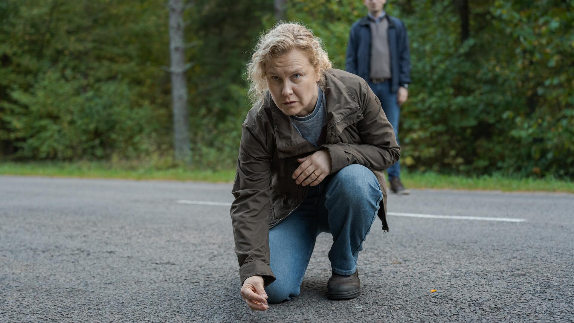 "Wolfssommer – Blutige Spuren - Die Spur der Wölfe": Hannah (Eva Melander) kniet auf einer Straße. Sie hebt etwas mit zwei Fingern auf. Vor ihr auf der Fahrbahn sind kleine Gegenstände zu sehen. Im Hintergrund steht Gordon (Hannes Fohlin) vor einem Waldstück.