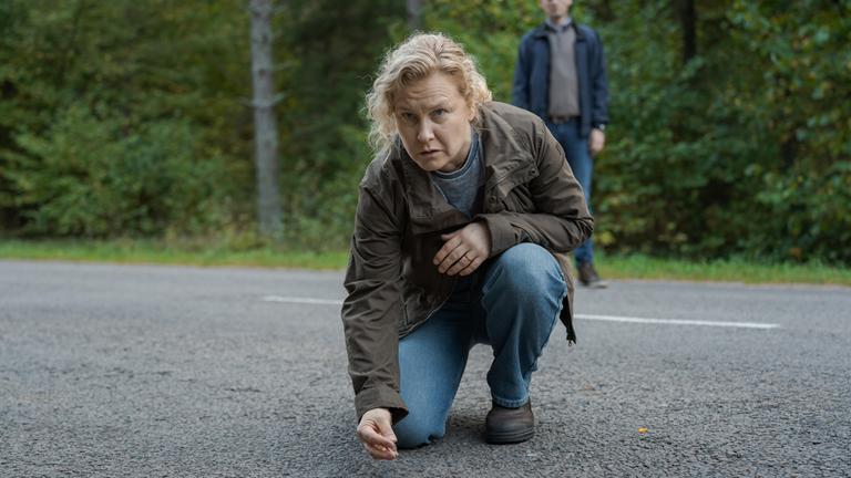 "Wolfssommer – Blutige Spuren - Die Spur der Wölfe": Hannah (Eva Melander) kniet auf einer Straße. Sie hebt etwas mit zwei Fingern auf. Vor ihr auf der Fahrbahn sind kleine Gegenstände zu sehen. Im Hintergrund steht Gordon (Hannes Fohlin) vor einem Waldstück.