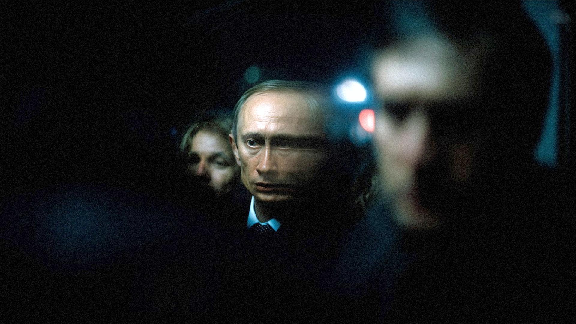 "Terra X History: Der Pate von St. Petersburg": Spielszene, Nachtaufnahme: Neben dem Kopf von Wladimir Putin (Marius Čizauskas) befinden sich zwei weitere Köpfe, die nur andeutungsweise, unscharf zu erkennen sind. Im Hintergrund leuchtet ein Licht.