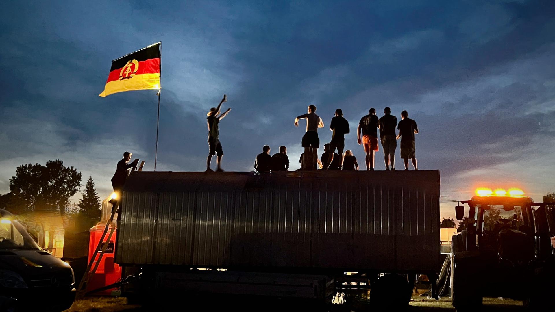 "Einigkeit, Verdruss und Freiheit - Wo stehen wir im Osten?": Menschen stehen im Abendlicht auf einem Anhänger neben ihnen weht die DDR-Fahne.