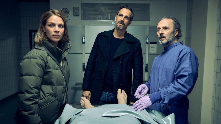 "Vogelfrei – Ein Schwarzwaldkrimi - Zwischenzeit": Maris (Jessica Schwarz), Konrad Diener (Max von Thun) und Dr. Stefan Zabel (Robert Schupp) stehen zusammen im Leichenschauhaus hinter einer Leiche.