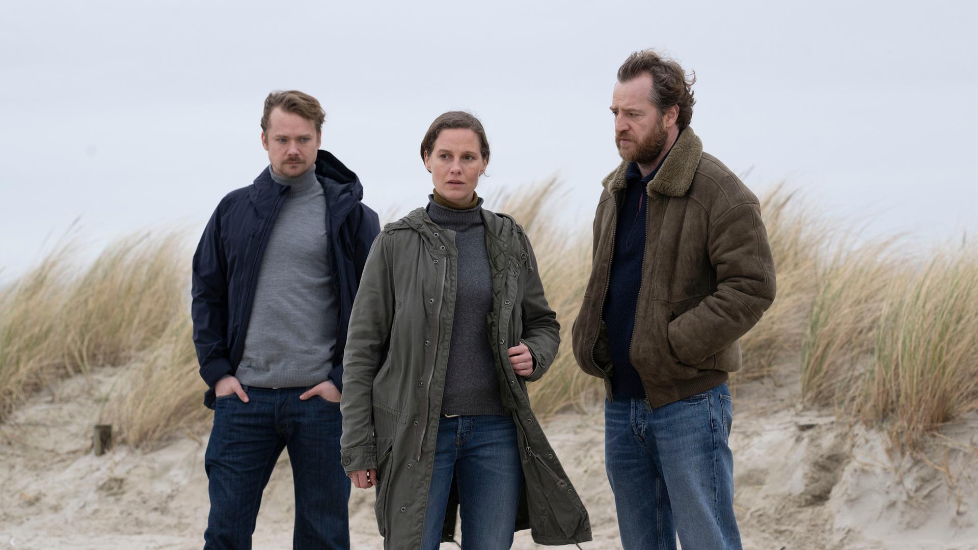 Kommissarin Ann Kathrin Klaasen (Picco von Groote) steht zusammen mit ihrem Kollegen Frank Weller (Tom Radisch) und Dr. Bernhard Sommerfeldt (Sven Schelker) am Rande einer Düne. Jeder hat einen nachdenklichen und ernsten Blick.