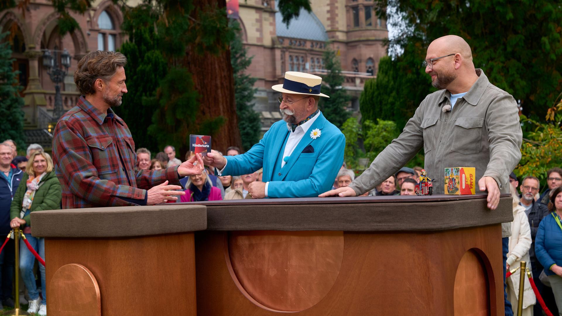 "Bares für Rares XXL – Einmal um die Welt": Vor Schloss Drachenburg: Benjamin Sadler, Horst Lichter und Sven Deutschmanek stehen hinter dem Expertisentisch. Horst Lichter überreicht Benjamin Sadler die Händlerkarte. Im Hintergrund stehen Zuschauer.