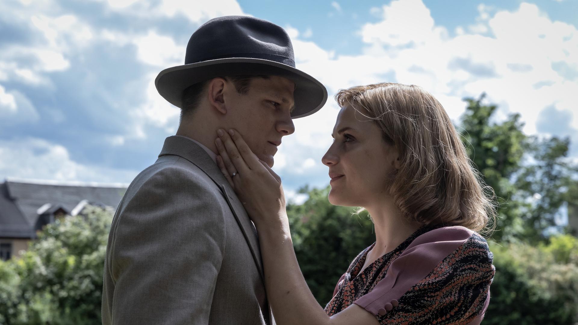 "Die Mittagsfrau": Helene (Mala Emde) steht mit Wilhelm (Max von der Groeben) auf einer Wiese und sie kommen sich näher. Im Hintergrund stehen Büsche vor einem Haus.