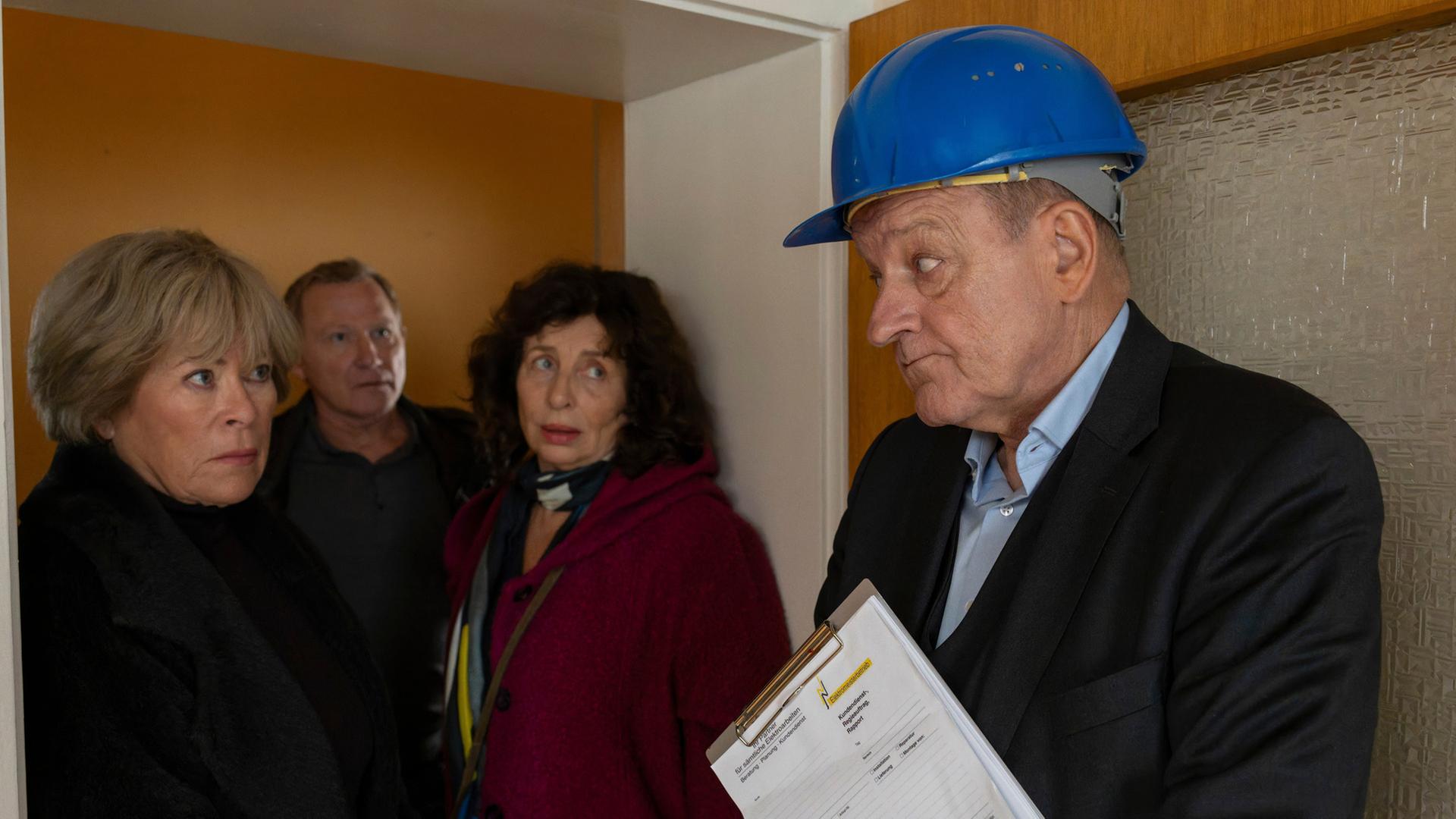 "Wilsberg - Mogelpackung": Wilsberg (Leonard Lansink), Anna Springer (Rita Russek), Overbeck (Roland Jankowsky) und Janne Becker (Teresa Harder) stehen gemeinsam im Türrahmen einer Wohnung. Wilsberg trägt einen blauen Baustellenhelm und hält ein Klemmbrett mit einem weißen Zettel in der Hand. Die anderen drei Personen blicken fragend in seine Richtung.