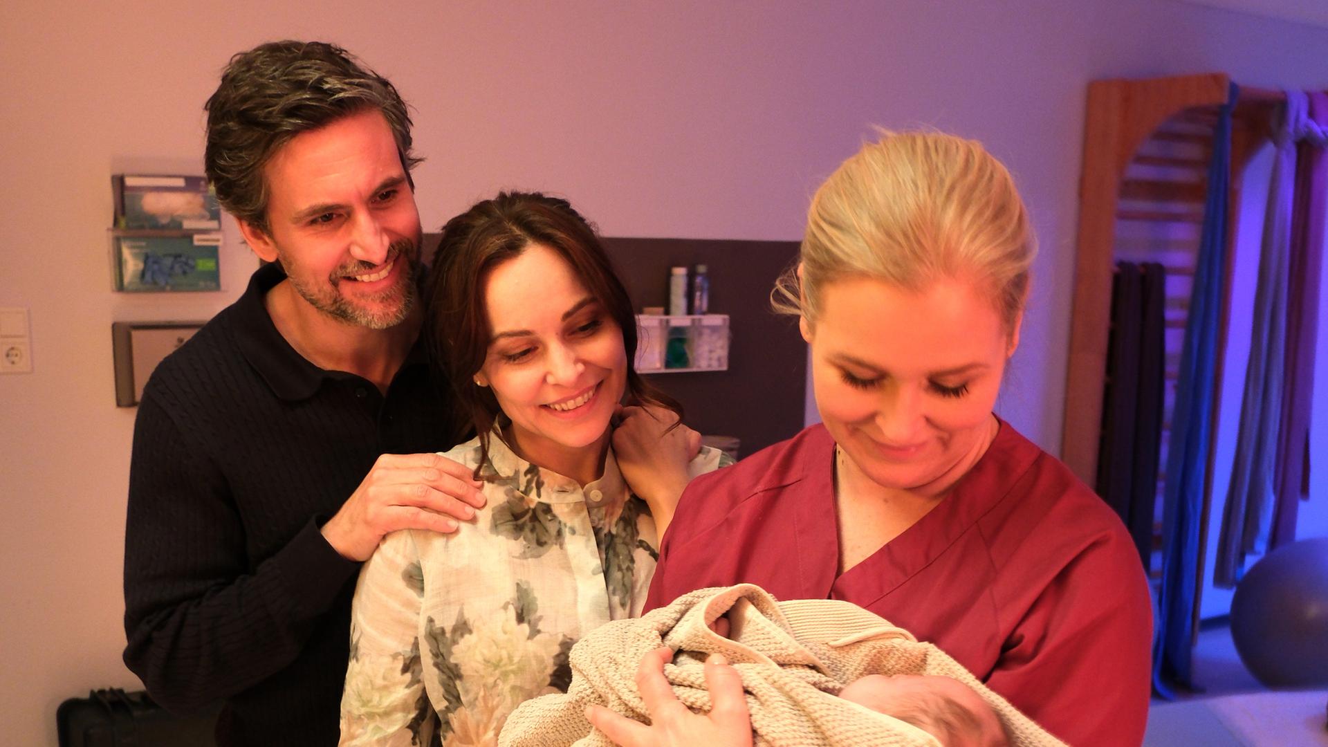 "Lena Lorenz - Muttergefühle": In einem Kreißsaal hält Lena Lorenz (Judith Hoersch) ein Baby im Arm, das in eine Decke gewickelt ist und betrachtet es lächelnd. Jan (Tom Beck) und Katina Wolff (Mina Tander) stehen mit strahlendem Lächeln daneben und schauen das Baby freudig an.