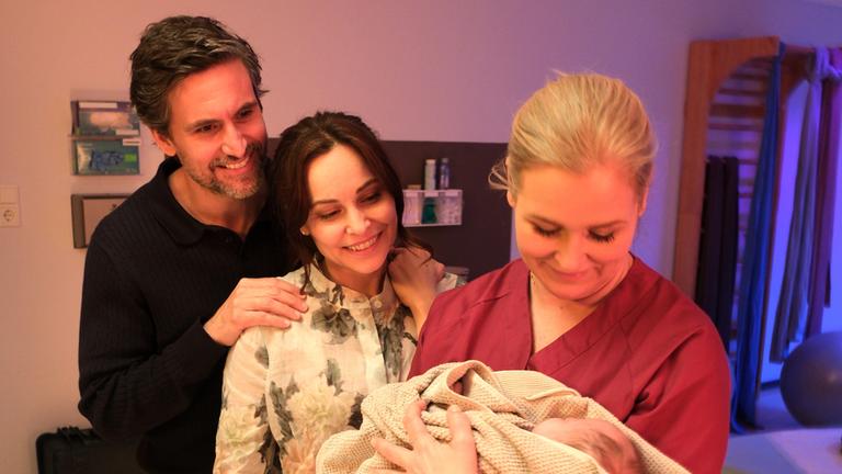 "Lena Lorenz - Muttergefühle": In einem Kreißsaal hält Lena Lorenz (Judith Hoersch) ein Baby im Arm, das in eine Decke gewickelt ist und betrachtet es lächelnd. Jan (Tom Beck) und Katina Wolff (Mina Tander) stehen mit strahlendem Lächeln daneben und schauen das Baby freudig an.