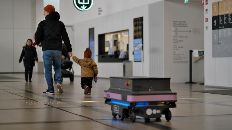 "plan b: Digitale Kollegen im Einsatz - Wie KI und Robotik unsere Jobs verbessern": In einem großen, hellen Flur in Helsinkis neuer Bücherei "Oodi" fährt ein Roboter an einer schwarzen Markierung entlang. Neben ihm laufen einige Personen.