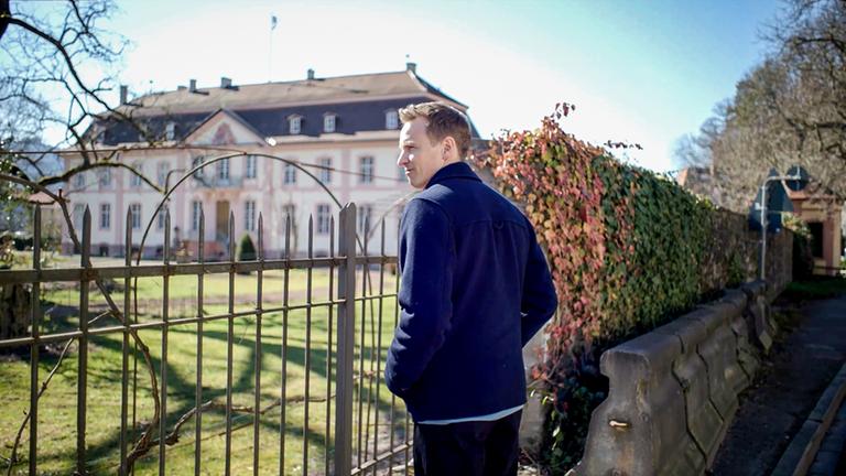 "Die geheime Welt des Adels - mit Jochen Breyer": Jochen Breyer blickt über einen Zaun, dahinter liegt ein Schloss im Sonnenschein.