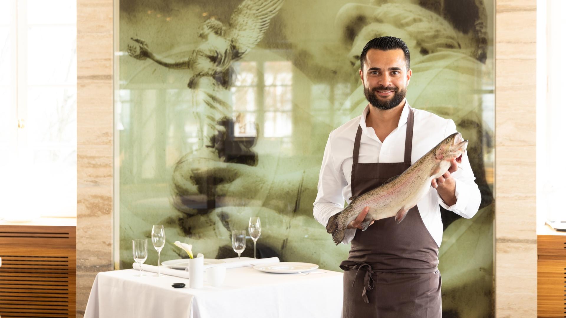  "ZDF.reportage: Essen - Hauptsache lecker": Küchenchef Dennis Kuckuck hält einen großen Fisch in der Hand und lächelt in die Kamera. Er trägt eine Schürze. Im Hintergrund ist der schick gedeckte Tisch eines Restaurants zu sehen.