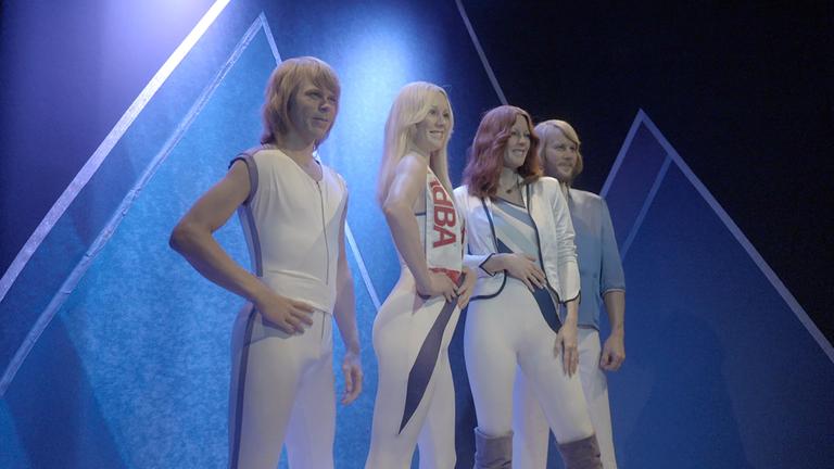 "Metropolen des Pop: Stockholm": Die vier Mitglieder der Band ABBA als Wachsfiguren in eng anliegenden, weißen Kostümen stehen nebeneinander unter blauem Licht und Nebel; im Hintergrund leuchten geometrische Rahmenelemente.