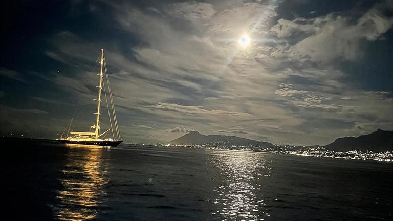 "Terra X: Das Drama der Superyacht Bayesian - Warum sinken Schiffe?": Die beleuchtete Segelyacht "Bayesian" liegt auf dem Meer vor bergiger Kulisse bei Mondschein.