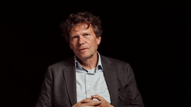 "Zwischen uns die Wahrheit": Porträtaufnahme: Hartmut Rosa sitzt vor dunklem Hintergrund. Sein Blick ist leicht zur Seite gerichtet, die Hände sind vor dem Körper gefaltet.