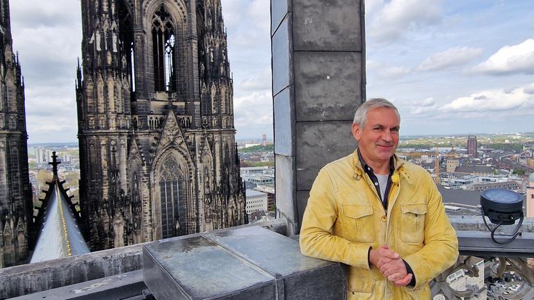Wladimir Kaminer auf einer Aussichtsplattform des Kölner Doms mit Blick auf einen der Türme und die Stadt Köln im Hintergrund.