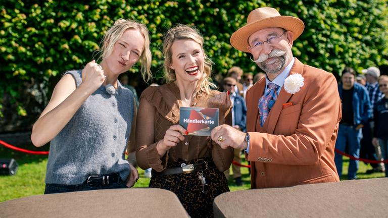 "Bares für Rares XXL": Leonie Bartsch, Linn Schütze, und Horst Lichter stehen hinter dem Expertisenpult und lächeln in die Kamera. Horst Lichter überreicht Linn Schütze die Händlerkarte. Im Hintergrund steht Publikum.