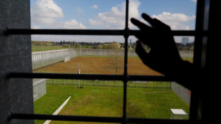 "Im Gefängnis: Das Prinzip Hoffnung": Eine Hand fasst an ein vergittertes Fenster. Hinter dem Fenster liegt ein großer Platz mit Rasen, der von einem hohen Zaun umfasst ist.