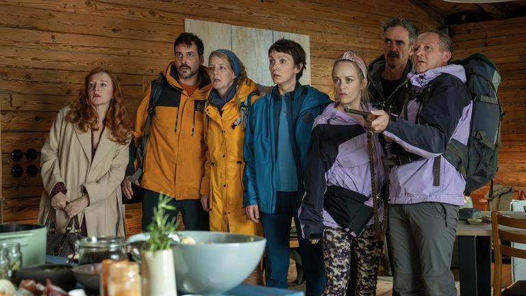 "Nix ist fix": Nebeneinander aufgereiht steht die Freundesclique vor einer hölzernen Wand im Chalet. Ines (Brigitte Hobmeier), Harry (Marcel Mohab), Sanne (Katrin Wichmann), Juliane (Julia Koschitz), Edwin (Felix Hellmann), Paula (Friederike Kempter) und Pascal (Maximilian Brückner) blicken mit entsetzten Gesichtern zur Seite. Pascal hält ein Gewehr in den Händen.