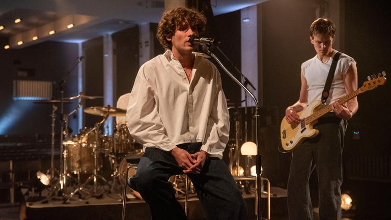"Pop Around@Bauhaus: Jeremias": Zwei Musiker der Band Jeremias während eines Auftritts auf einer Bühne. Eine Person sitzt auf einem Hocker und singt in ein Mikrofon. Eine weitere Person steht etwas weiter hinten und spielt auf einer E-Gitarre.