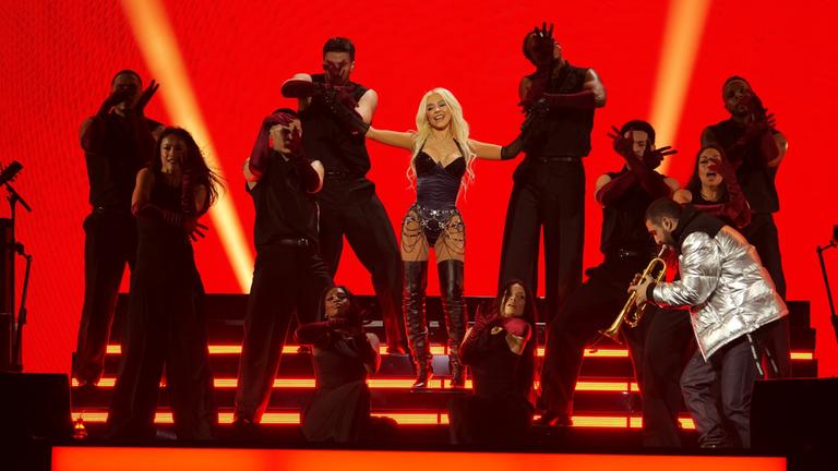 "Christina Aguilera, Stray Kids, Davido, Future, G-Dragon, A$AP Rocky & Friends: Le Gala des Pièces Jaunes 2026": Sängerin Christina Aguilera steht mittig auf einer Showtreppe. Das Scheinwerfer-Licht ist auf sie gerichtet. Um sie herum befinden sich zahlreiche weitere Personen, die tanzen oder singen. Der Hintergrund ist in einem orange-roten Farbton gehalten.