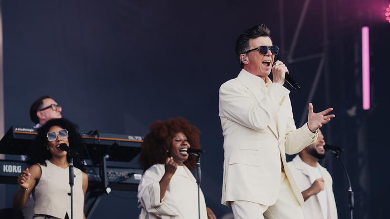 "Isle of Wight Festival 2026": Rick Astley steht auf einer Bühne und singt. Hinter ihm stehen zwei Background-Sängerinnen und ein -Sänger sowie ein Musiker an einem E-Piano.
