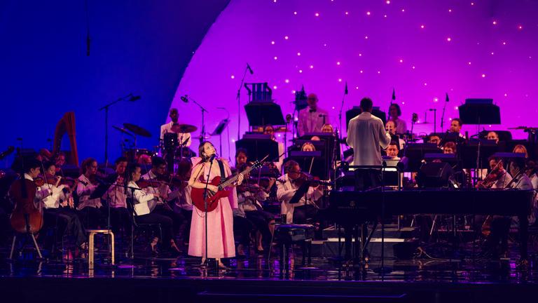 "Laufey: A Night at the Symphony": Die Musikerin Laufey während eines Auftritts auf einer Bühne. Sie hält eine Gitarre in der Hand und singt in ein Mikrofon. Hinter ihr die Musiker eines Orchesters und der Dirigent. Der Hintergrund zeigt eine lila-pinke Lichtstimmung