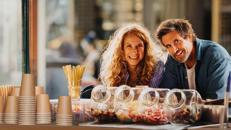 "Eine Wingwoman verliebt sich nicht": Caro Sander (Chiara Schoras) und Marc Ahmadi (Tom Beck) stehen nebeneinander an einem Kiosk. Vor ihnen stehen Süßigkeiten in Glasbehältern und Pappbecher. Beide schauen lächelnd nach vorne.