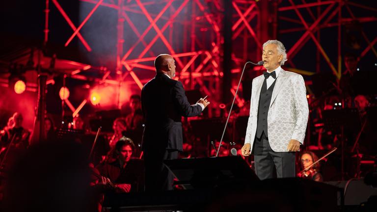 "Andrea Bocelli: Romanza": Andrea Bocelli steht auf einer Bühne und singt. Links neben ihm steht der Dirigent und im Hintergrund sitzen die Musiker des Orchesters. Die Bühne ist in rotes Licht getaucht.