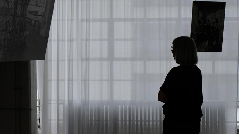 "Der Kampf um die Kultur – Ostdeutschland vor der Wahl": Eine Frau steht vor einer großen Fensterfront, davor ist ein transparenter, weißer Vorhang. Sie schaut auf eine Fotografie, die man links oben erkennen kann.