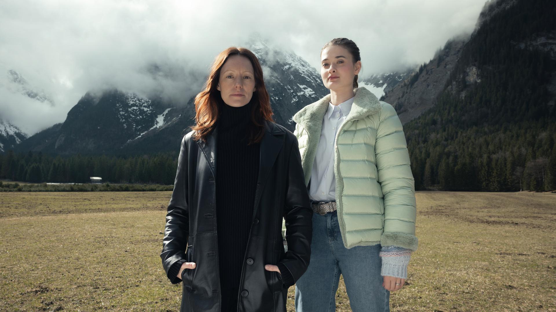 “Der Garmisch-Krimi - Wolfsmord“: Ira Zach (Lavinia Wilson) und Daphne Meindl (Philine Schmölzer) stehen auf einer Wiese in Garmisch vor hohen Bergen, deren Gipfel der Nebel bedeckt.