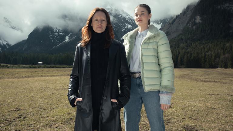 “Der Garmisch-Krimi - Wolfsmord“: Ira Zach (Lavinia Wilson) und Daphne Meindl (Philine Schmölzer) stehen auf einer Wiese in Garmisch vor hohen Bergen, deren Gipfel der Nebel bedeckt.