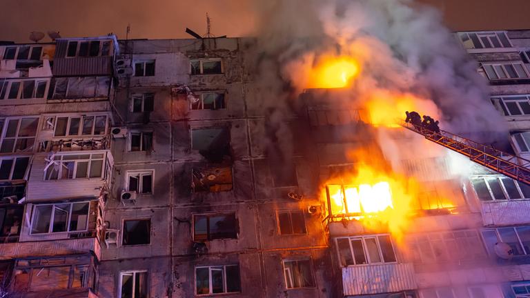 "Terra X History: Putins Blutspur - Chronik eines Krieges (4)": Untersicht: In einem mehrstöckigen Haus sind nachts in drei Stockwerken lodernde Brände. Eine Feuerwehrleiter ragt an der Fassade empor, zwei Feuerwehrmänner stehen darauf. Rauch steigt auf.