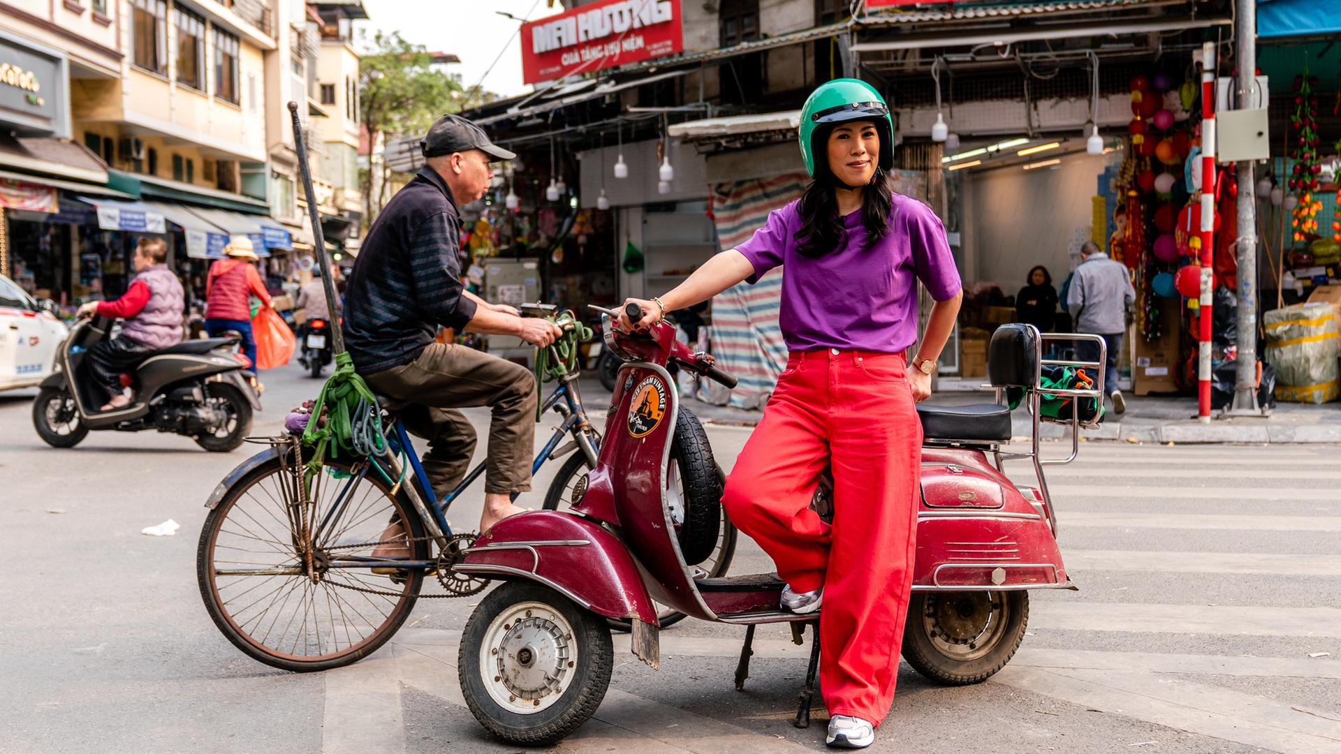 "Terra X: Vietnam - Eine Reise mit Mai Thi Nguyen-Kim": Mai lehnt sich an einen roten Moped, mitten auf der Straße. Im Hintergrund befinden sich kleine Märkte, Straßen und Wege sind belebt.