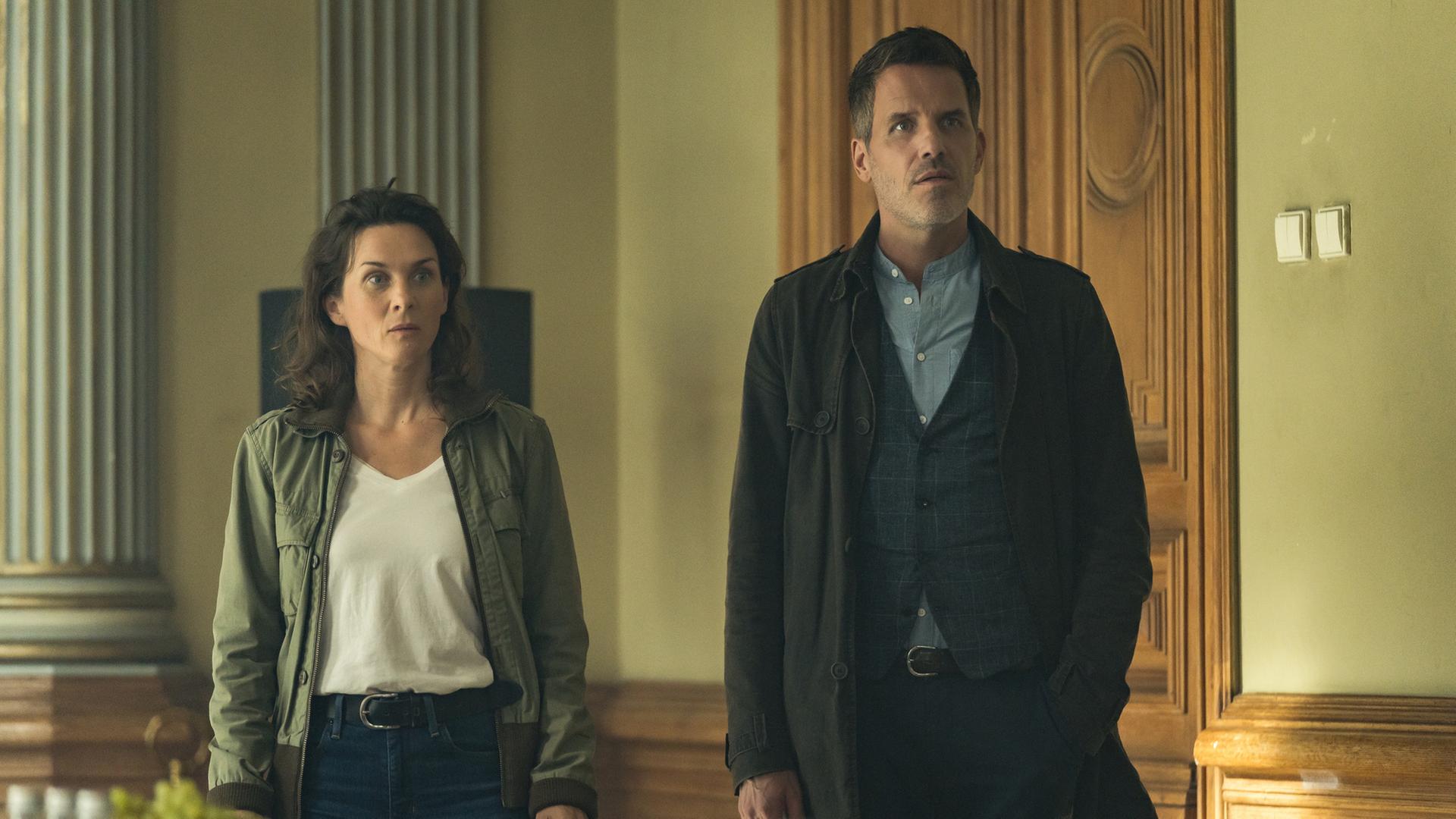 Bildbeschreibung: "In ewiger Freundschaft (1) - Ein Taunuskrimi": Mit offenen Blicken steht Pia Sander (Kathrin von Steinburg) neben ihrem Kollegen Oliver von Bodenstein (Tim Bergmann) in einem Raum mit antiken Säulen vor einer hohen, aufwendig gestalteten Holztür.