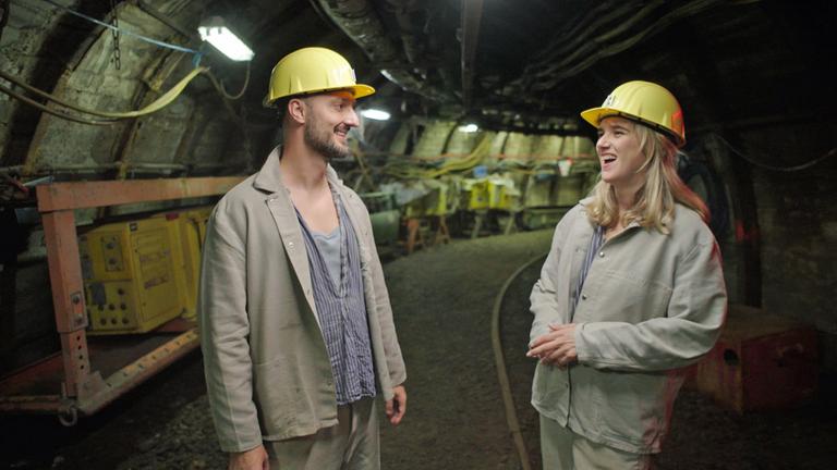 "WISO-Dokumentation: Die Welt in Geld - Kryptowährungen": Frederik Dürr und Katjana Gerz  in hellen Bergbauanzügen und mit gelben Schutzhelmen im Gespräch unter Tage. Im dunklen Hintergrund ist ein Gang erkennbar.