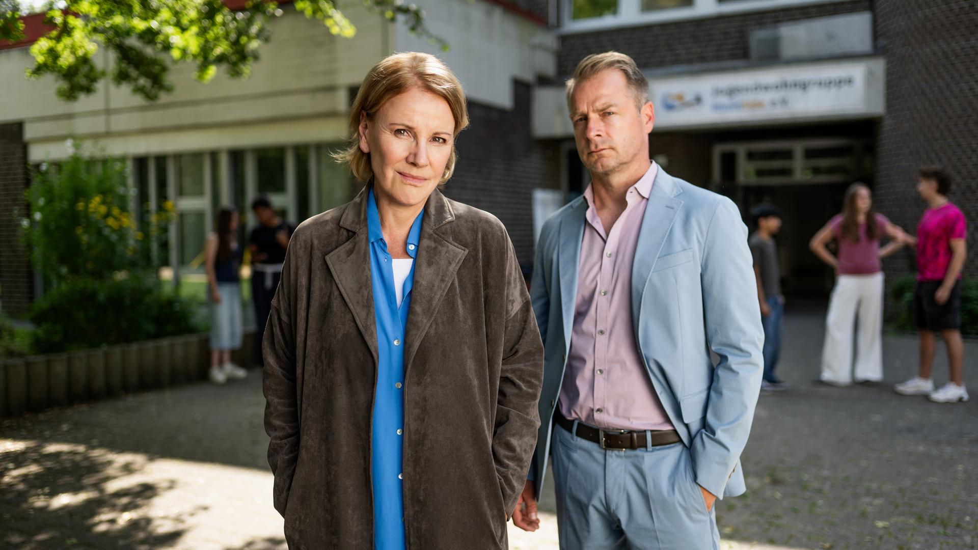 Bildbeschreibung: "Marie Brand und die verlorenen Kinder": Marie Brand (Mariele Millowitsch) steht zusammen mit Jürgen Simmel (Hinnerk Schönemann) vor dem Eingang eines Kinderhorts. Im Hintergrund stehen mehrere Jugendliche (Komparsen) vor den Eingangstüren.