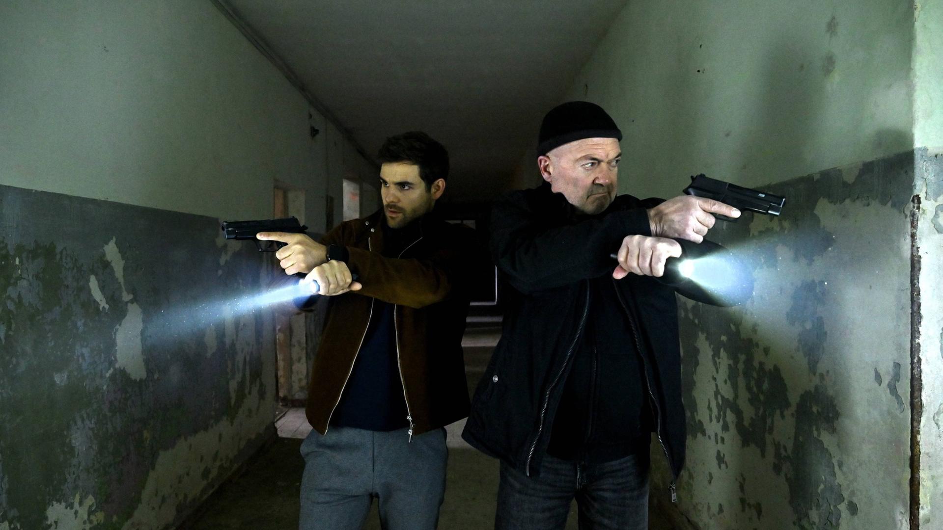 "Ein starkes Team - Das letzte Opfer": Nils Makowski (Lucas Reiber) und Otto Garber (Florian Martens) stehen in einem breiten dunklen Gang. Das Gebäude ist heruntergekommen. Beide haben ihre Waffe gezogen und leuchten mit Taschenlampen in die entgegengesetzte Richtung.