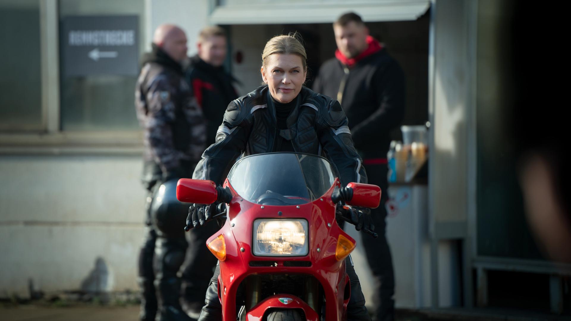 "Helen Dorn - Adrenalin": Helen Dorn sitzt auf einem Motorrad und fährt Richtung Kamera. Im Hintergrund, unscharf zu erkennen, stehen drei Männer vor einem Gebäude.