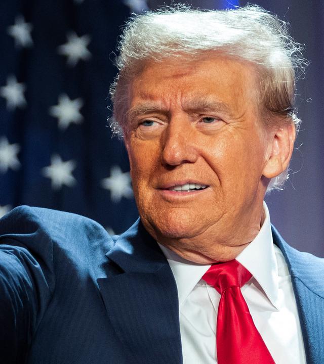 "Donald Trump - König im Weißen Haus?": Porträtaufnahme: Donald Trump steht im Anzug vor einer amerikanischen Flagge. Er zeigt mit dem rechten Arm und ausgestrecktem Zeigefinger auf etwas. Sein Blick folgt seinem Arm.