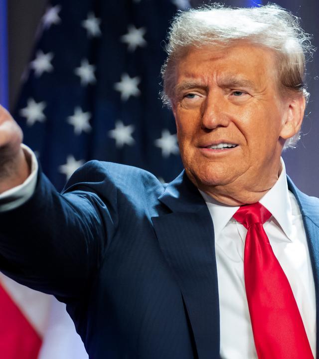 "Donald Trump - König im Weißen Haus?": Porträtaufnahme: Donald Trump steht im Anzug vor einer amerikanischen Flagge. Er zeigt mit dem rechten Arm und ausgestrecktem Zeigefinger auf etwas. Sein Blick folgt seinem Arm.