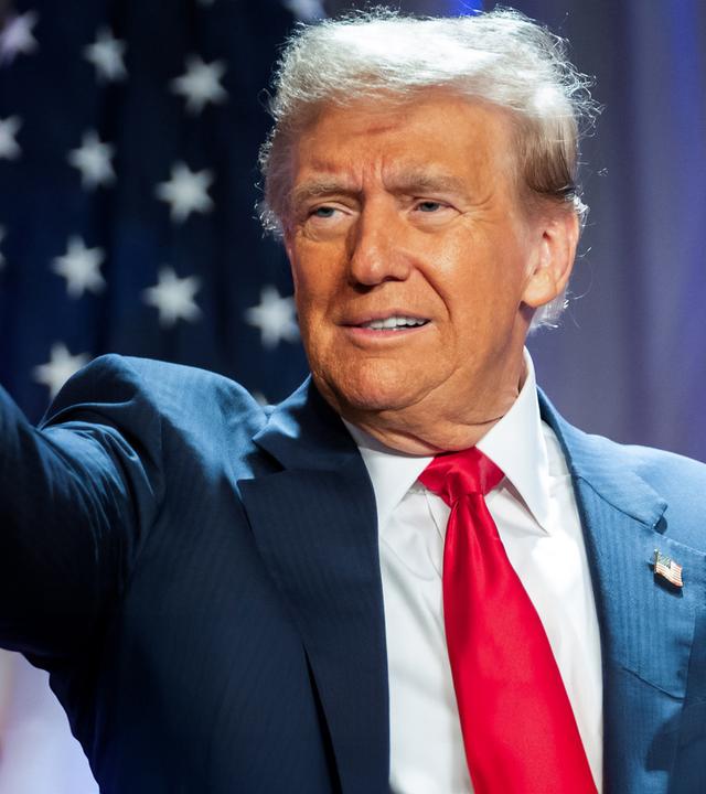 "Donald Trump - König im Weißen Haus?": Porträtaufnahme: Donald Trump steht im Anzug vor einer amerikanischen Flagge. Er zeigt mit dem rechten Arm und ausgestrecktem Zeigefinger auf etwas. Sein Blick folgt seinem Arm.