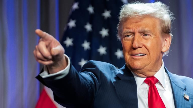 "Donald Trump - König im Weißen Haus?": Porträtaufnahme: Donald Trump steht im Anzug vor einer amerikanischen Flagge. Er zeigt mit dem rechten Arm und ausgestrecktem Zeigefinger auf etwas. Sein Blick folgt seinem Arm.