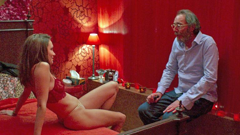 "La Maison – Haus der Lust": Emma (Ana Girardot) sitzt in einem rot beleuchtenden Zimmer in BH und Slip zurückgelehnt auf seinem Bett mit roter Decke. Sie blickt Hermann (Philippe Rebbot) lächelnd an, der ihr angezogen und mit ratloser Mine vor roten Vorhängen gegenübersitzt.