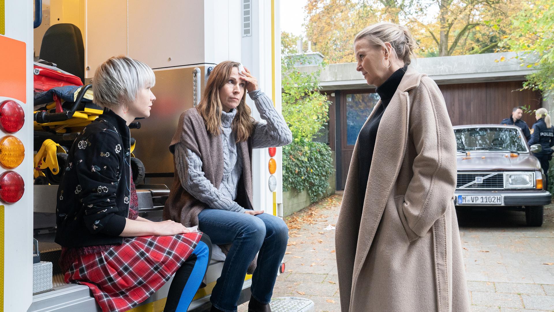 "Die Chefin - Schattenkrieger": Sabine Hübner (Doris Schretzmayer) sitzt mit ihrer Tochter Hanna Hübner (Emma Stratmann) auf den Stufen eines Rettungswagens. Sabine Hübner drückt sich ein Taschentuch an die Stirn. Vor ihnen steht Vera Lanz (Katharina Böhm). Im Hintergrund liegt die Einfahrt zu dem Haus der Hübners, in der zwei weitere Polizisten (Komparsen) neben einem Auto stehen.