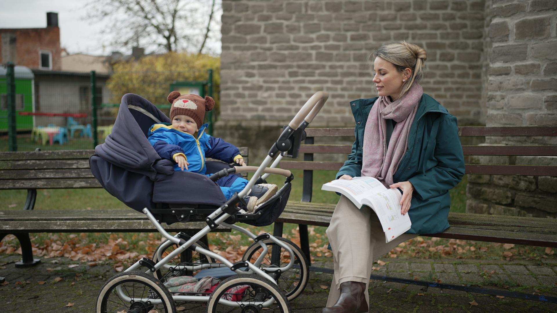 "37°: Arm, abhängig, abgehängt: Frauen und Geld": Carolin sitzt auf einer Bank im Freien, neben ihr Sohn Carl im Kinderwagen. Auf ihrem Schoß liegt ein Buch, das sie festhält. Sie schaut zum Kinderwagen.