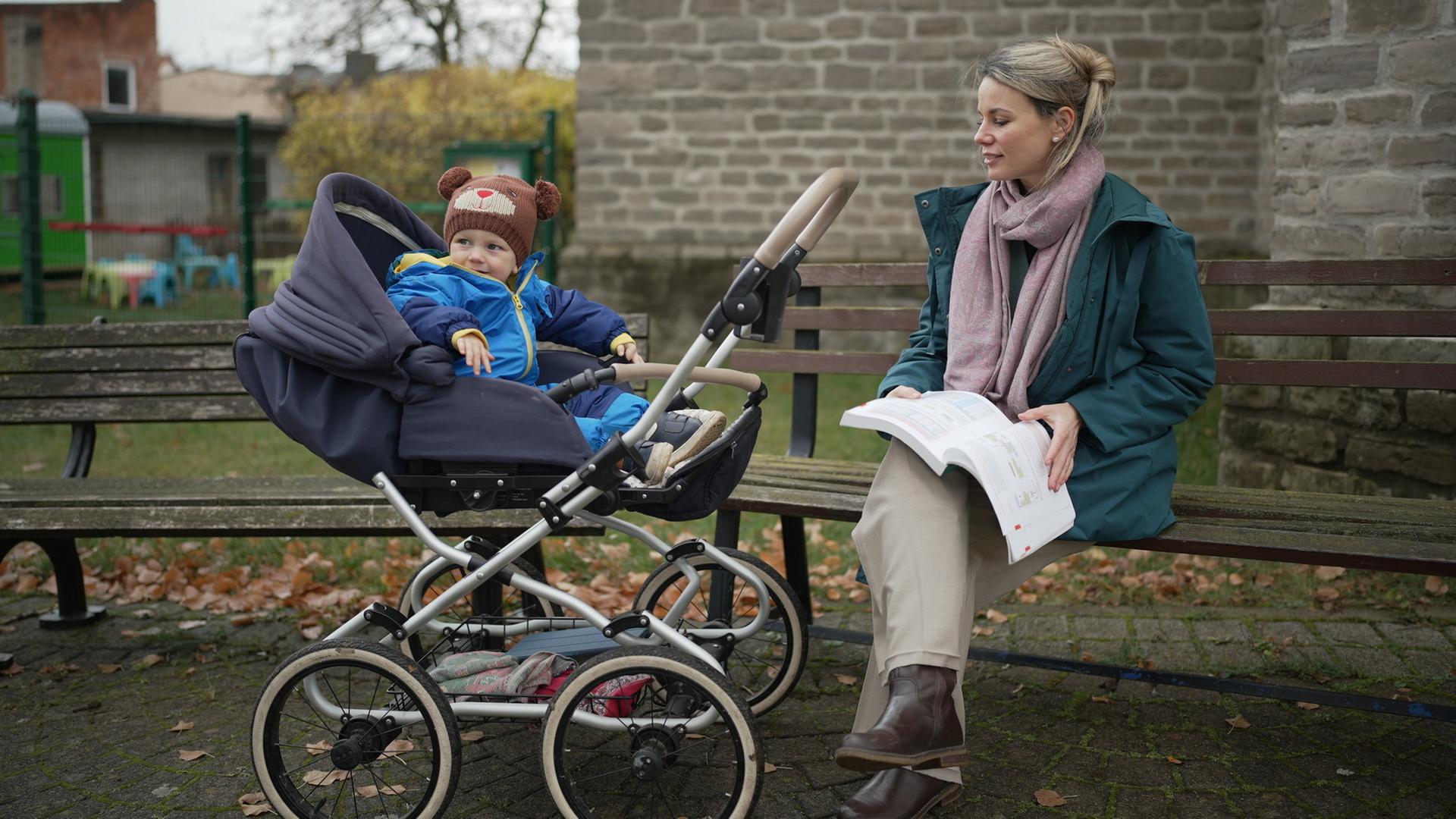 "37°: Arm, abhängig, abgehängt: Frauen und Geld": Carolin sitzt auf einer Bank im Freien, neben ihr Sohn Carl im Kinderwagen. Auf ihrem Schoß liegt ein Buch, das sie festhält. Sie schaut zum Kinderwagen.