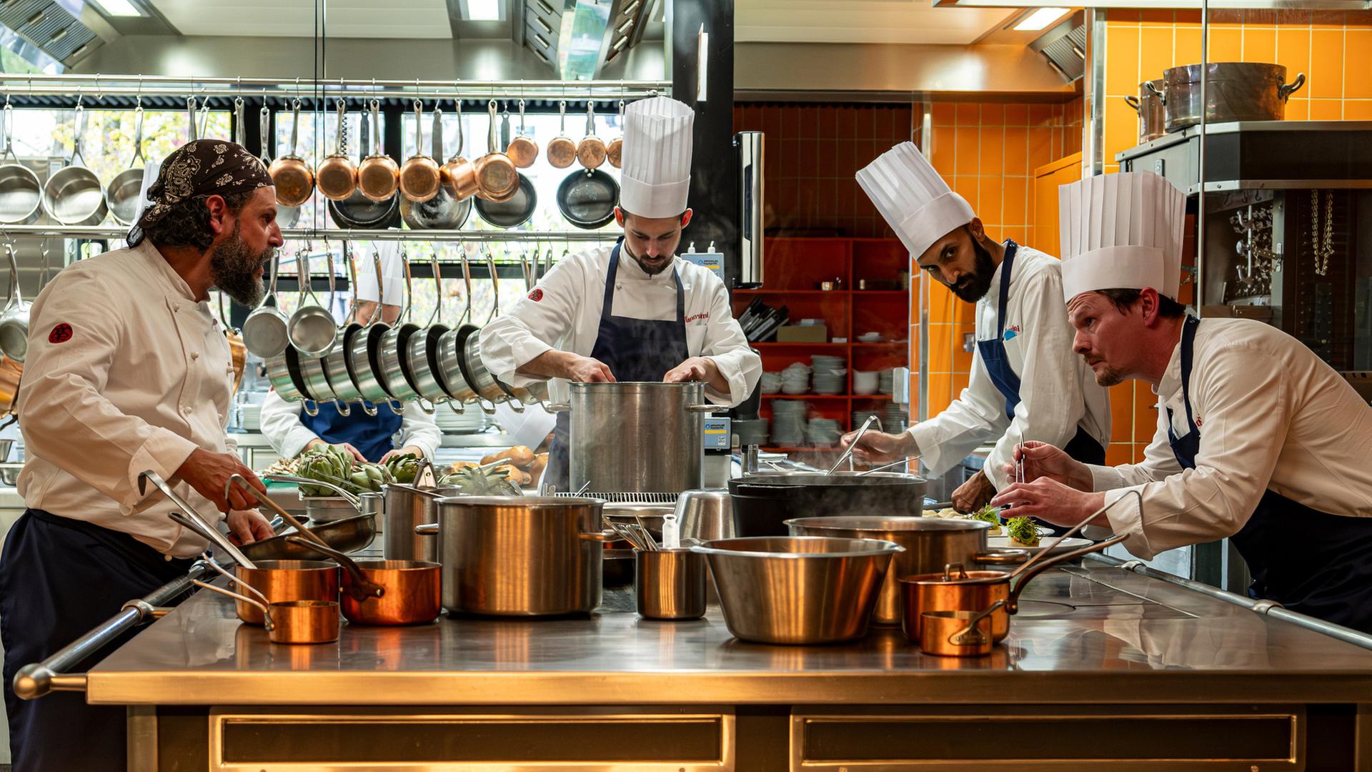 "München Mord - Im Zweifel für den Zweifel": Maximilian Dobek (Manuel Cortez) steht gemeinsam mit Oliver Gerg (Shenja Lacher) und zwei Beiköchen (Komparsen) in einer professionellen Küche. Alle tragen weiße Kochjacken und klassische Kochmützen. Vor ihnen erstreckt sich ein großer Arbeitstisch, auf dem zahlreiche Töpfe und Pfannen dampfen und brutzeln – jeder ist konzentriert bei der Sache. Im Hintergrund hängen Küchenutensilien ordentlich an Haken.