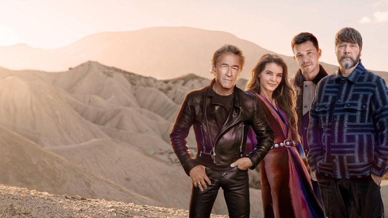 "Song Trip: Rea Garvey x Sápmi": Peter Maffay, Yvonne Catterfeld, Nico Santos und Rea Garvey stehen nebeneinander vor einer offenen, sandfarbenen Hügellandschaft. Die Umgebung besteht aus erodierten Hügeln ohne Vegetation. Das Licht der tiefstehenden Sonne taucht die Szene in warme, goldene Töne und erzeugt eine ruhige, atmosphärische Stimmung.